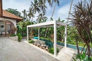 A pool area with a pergola, palm trees, and a house with a tiled roof.