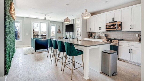 A modern kitchen with white cabinets, a central island with bar stools, a stainless steel refrigerator, and a large window providing natural light. - Harmony House #6–Roomy Retreat–Sleeps 12 (Nashville)