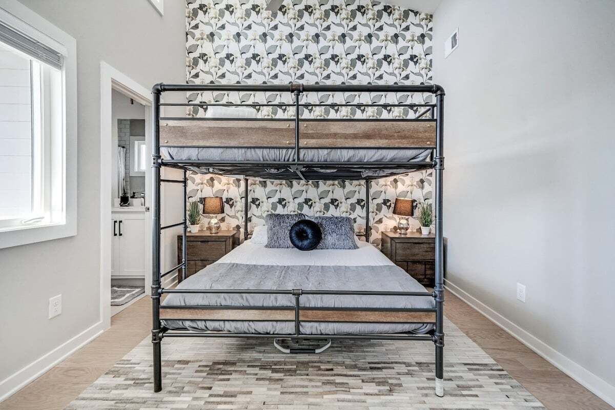A bunk bed with a wooden frame and grey bedding in a room with a patterned wallpaper and a small plant on the nightstand.