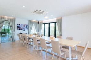 A spacious dining area with a long table and chairs, large windows with curtains, and a mounted television.