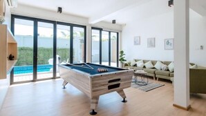 A modern living room with a pool table, large windows, and a green sofa. - Minimalist 4BR Private Cozy Pool Villa - VVP26.11 (Pattaya)