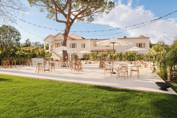 An outdoor patio with tables and chairs, surrounded by greenery and a large tree. - Auberge Tao (Biot)