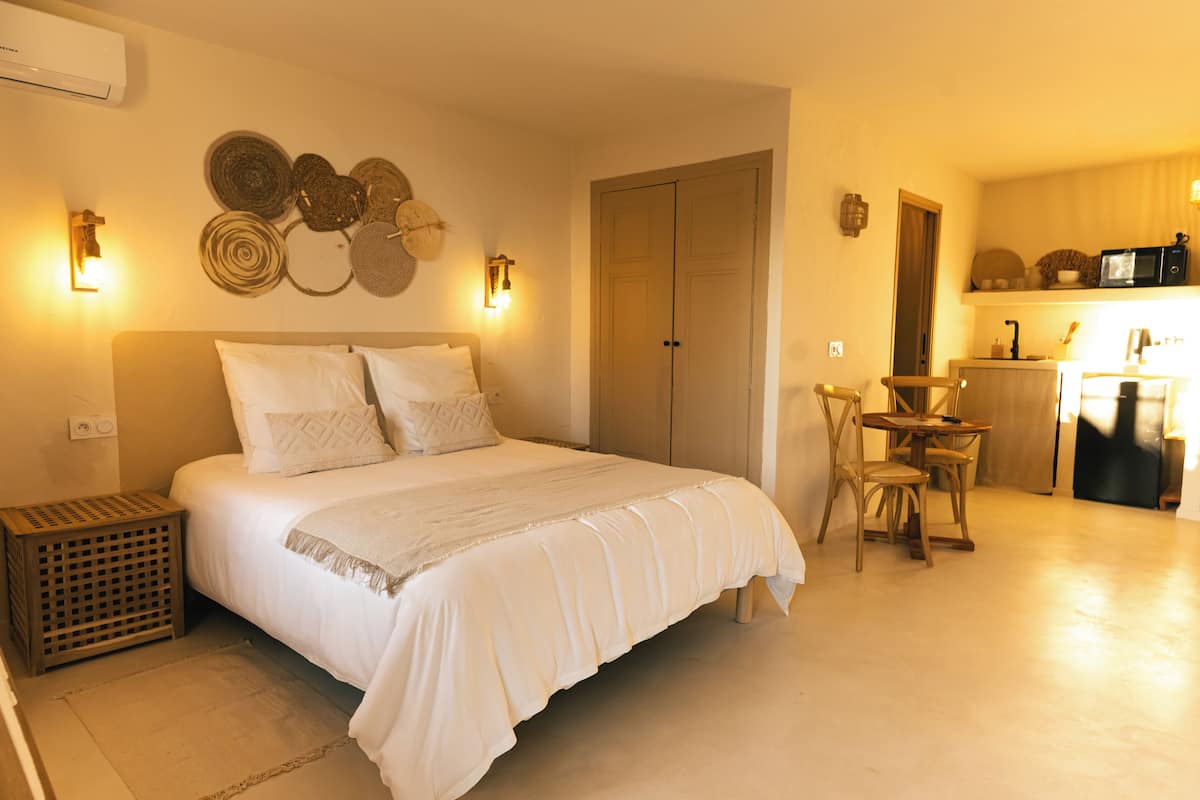 A bedroom with a bed, bedside table, and a small kitchen area with a microwave and a refrigerator.