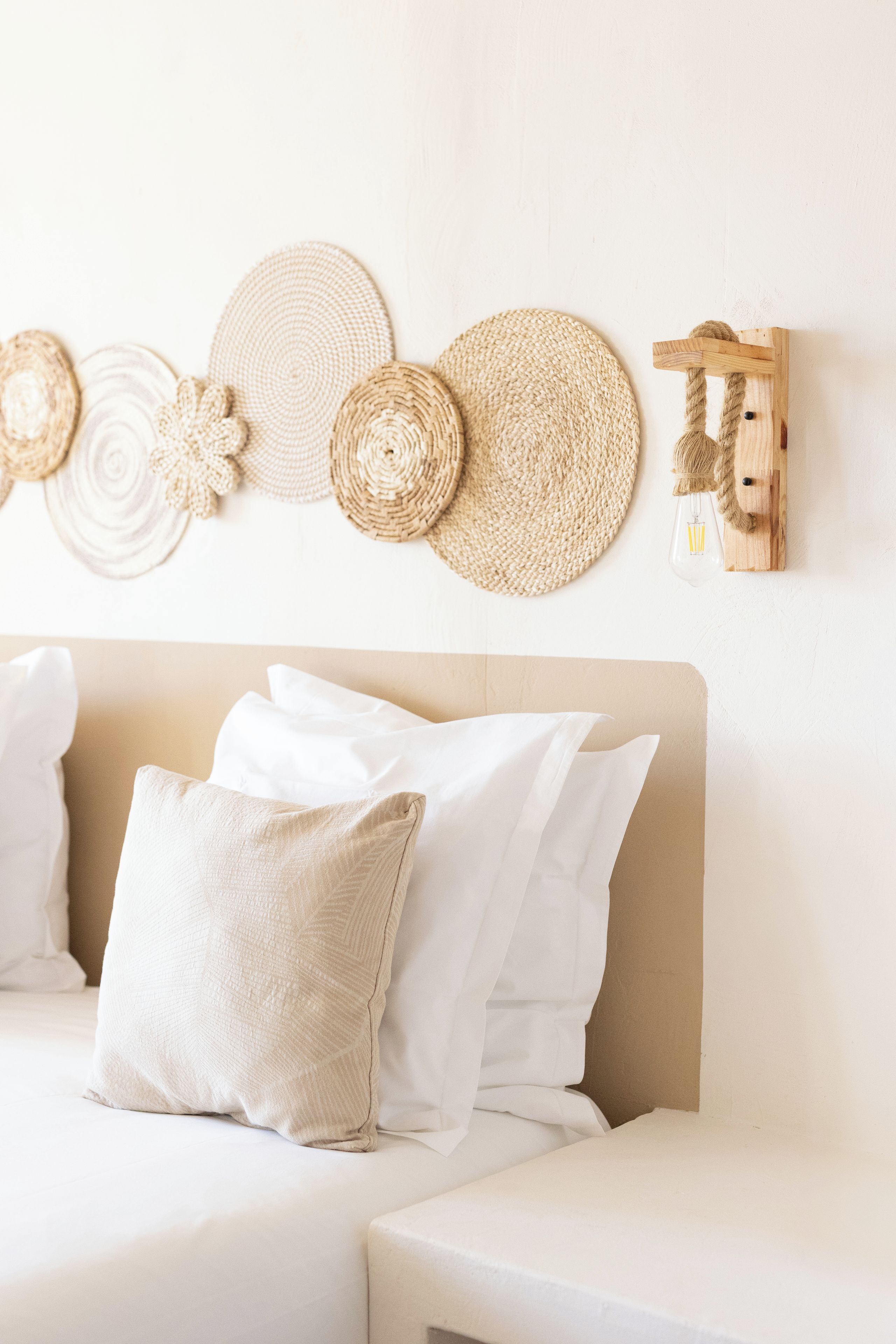Un lit avec une literie blanche et un oreiller beige, un mur orné d’une décoration murale tissée, et un luminaire en bois fixé au mur.