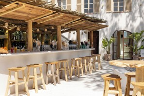 An outdoor bar area with wooden stools and tables, a bar counter with bottles and glasses, and a building with windows in the background. - Auberge Tao (Biot)