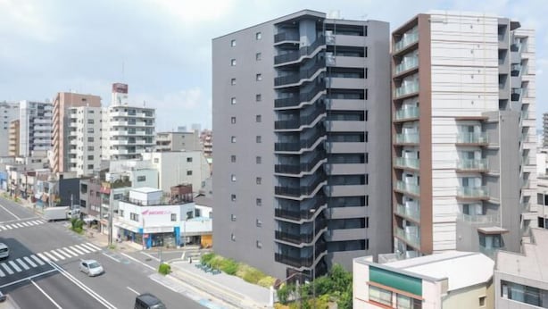A modern urban street with multi-story residential buildings, a crosswalk, and a storefront. - Grace Mukojima Room 701 (Tokyo)
