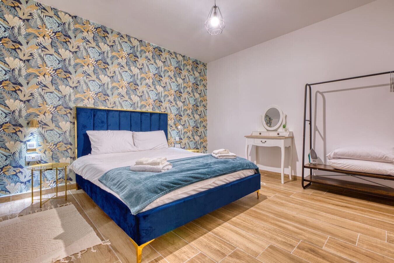 A bedroom with a blue bed, a white bedside table, a wooden floor, and a patterned wallpaper.