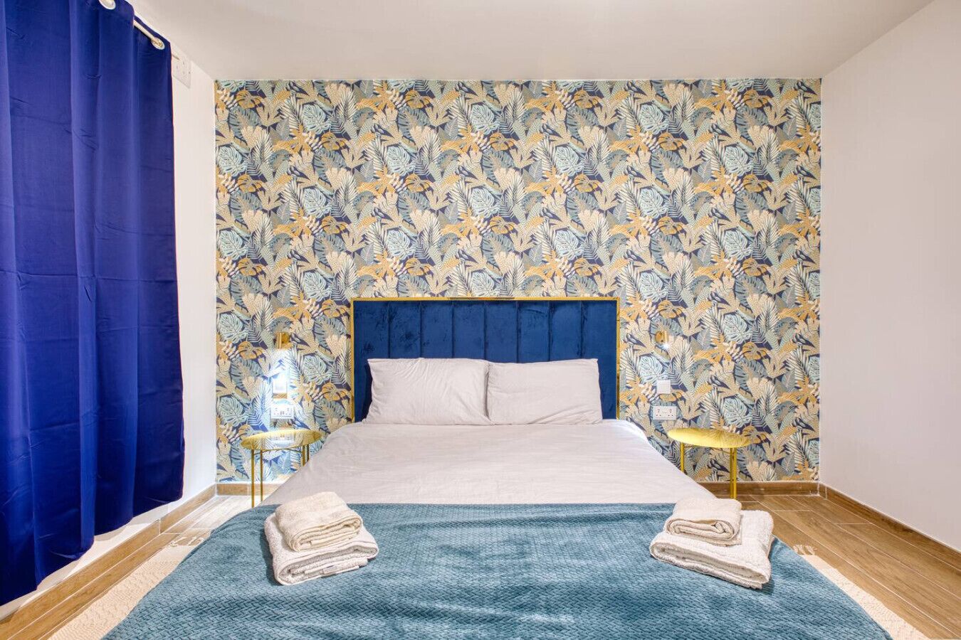 Une chambre à coucher avec un lit, un rideau bleu, un papier peint à motifs, une tête de lit bleue et deux tables de chevet avec des lampes.