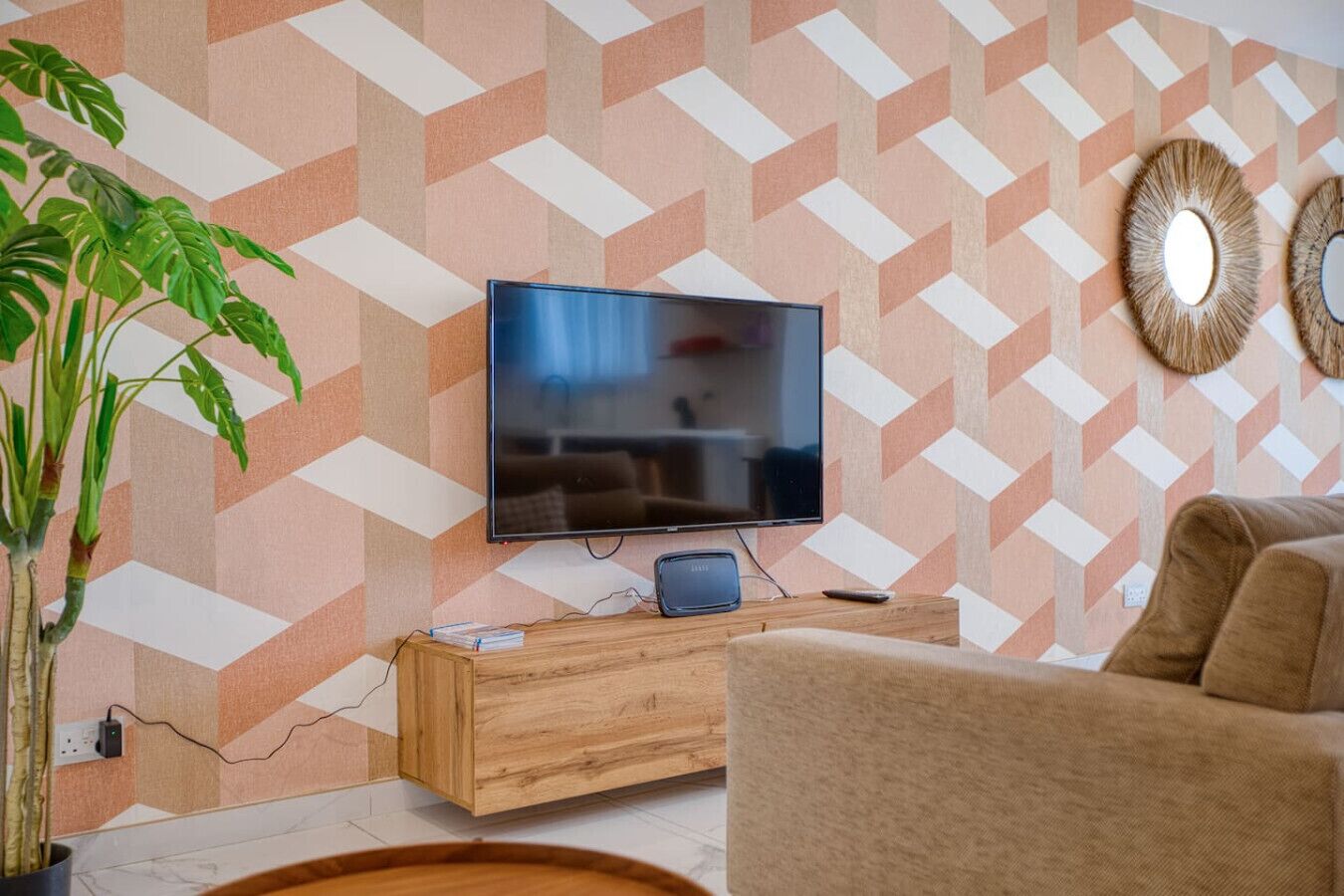 A living room with a flat-screen TV mounted on a wall with a geometric patterned wallpaper. There is a wooden console table in front of the TV, and a large potted plant to the left.