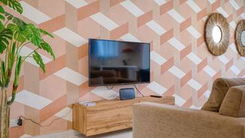 A living room with a flat-screen TV mounted on a wall with a geometric patterned wallpaper. There is a wooden console table in front of the TV, and a large potted plant to the left.
