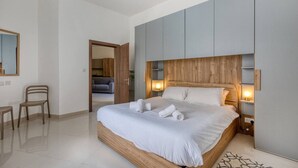 A modern bedroom with a large bed, wooden headboard, bedside tables with lamps, a chair, and a mirror on the wall. - Relaxing Xaghra Apartment With BBQ Amenities (Xaghra)