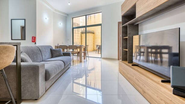 A modern living room with a grey sofa, a wooden TV stand, and a dining area with a table and chairs. - Relaxing Xaghra Apartment With BBQ Amenities (Xaghra)