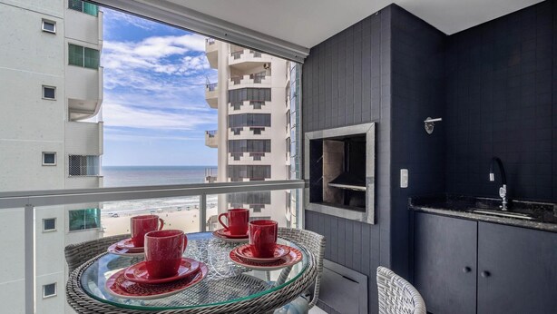 Un balcone con un tavolino in vetro e bicchieri rossi, con vista su una spiaggia e palazzi alti.