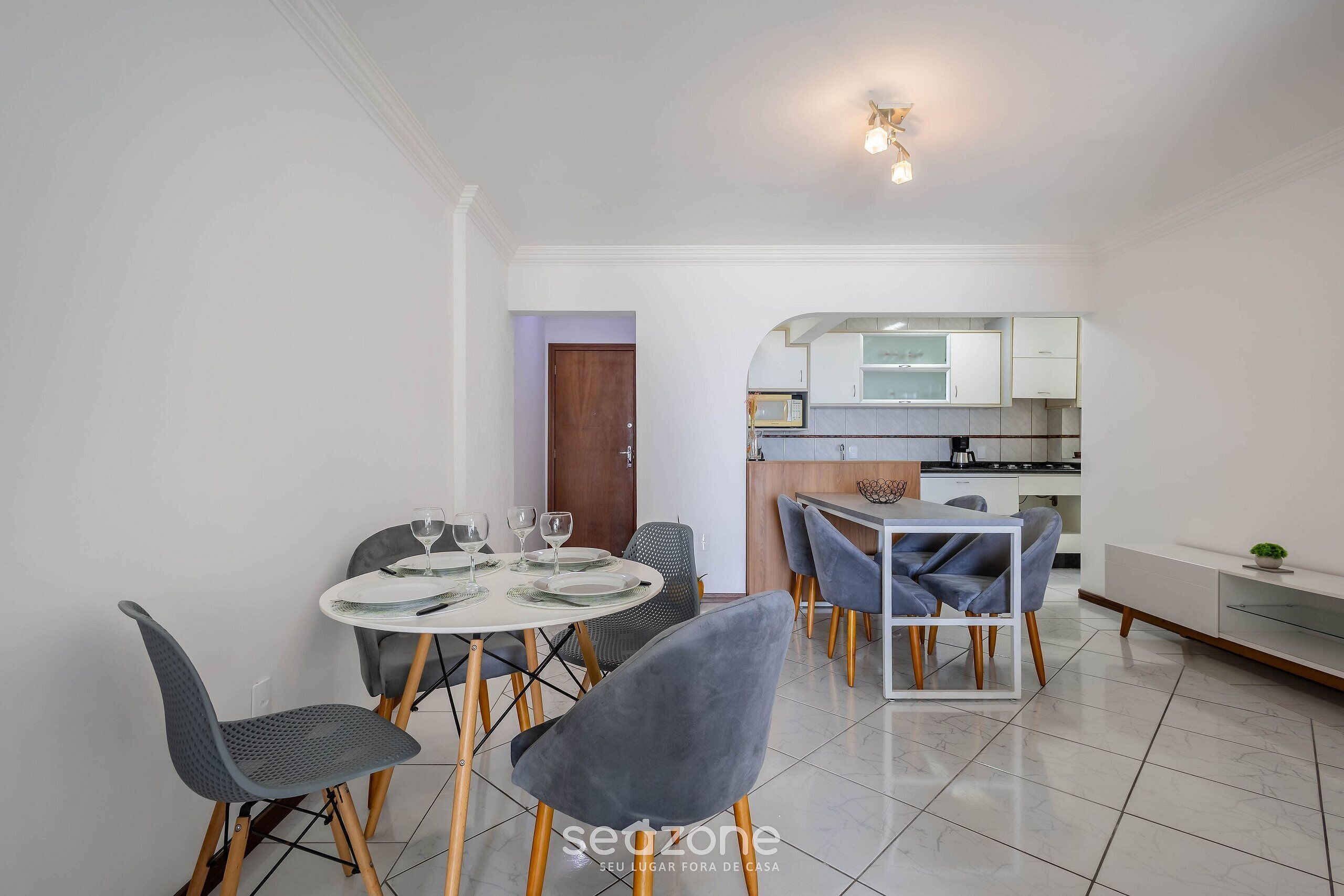 A modern dining area with a round table, grey upholstered chairs, and a kitchen in the background.