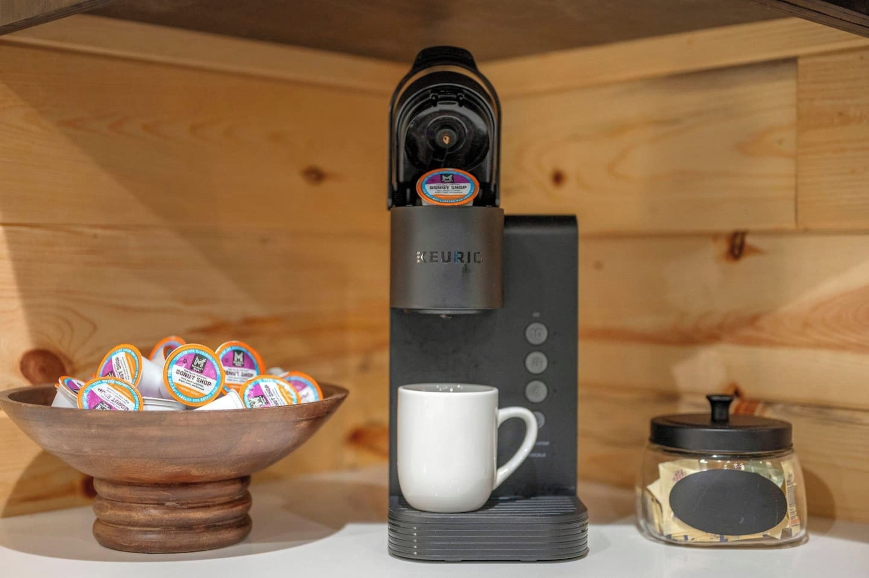 Una cafetera Keurig con una taza blanca, un bol de cápsulas de café y un tarro de café molido.