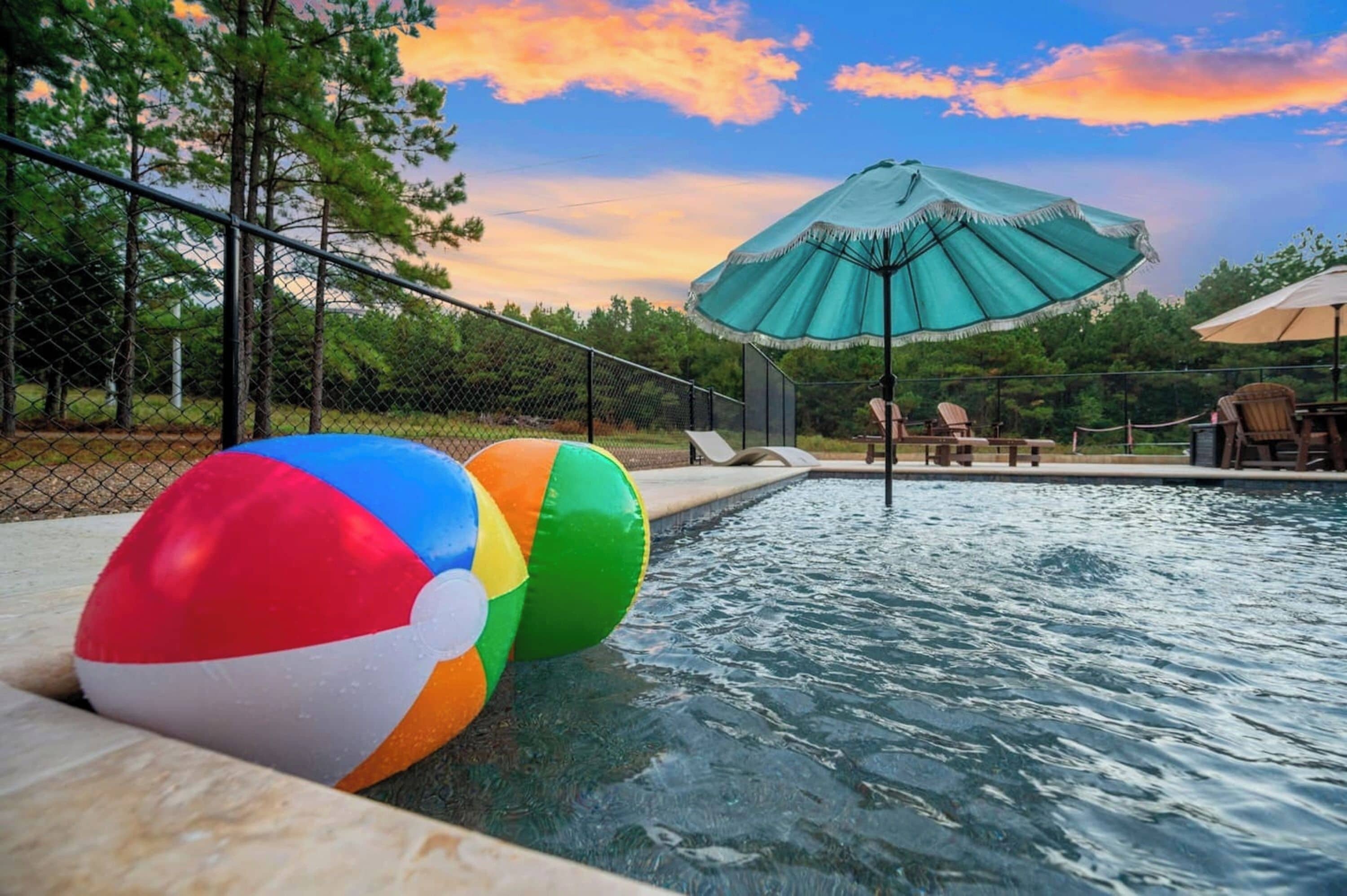 Una piscina con pelotas de playa, un paraguas verde y sillas de descanso.