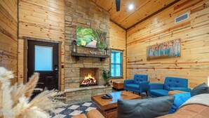A cozy cabin interior with a stone fireplace, blue upholstered chairs, and a checkered floor rug. - The Pickleball Pines (Broken Bow)