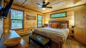 A wooden cabin bedroom with a bed, a bench, a television, and a ceiling fan. - The Pickleball Pines (Broken Bow)