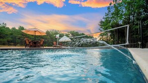 A swimming pool with a water feature, surrounded by trees and a sunset sky. - The Pickleball Pines (Broken Bow)