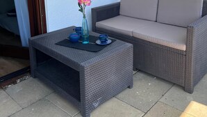 Un canapé en osier et une petite table sur laquelle se trouvent un vase et des tasses, le tout placé sur une terrasse.