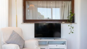 A living room with a flat-screen TV mounted on a white cabinet, a sofa with cushions, a floor lamp, and a window with curtains. - Rental in Rome San Pancrazio Penthouse (Rome)