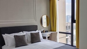 Habitación de hotel con cama, escritorio, silla, espejo redondo y ventana con vista a edificios.