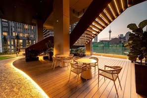 A rooftop terrace with wooden flooring, round tables, and chairs, overlooking a cityscape with a tower. - AXON Bukit Bintang Luxury Condo 3BED 2BATH (HIGH FLOOR) (Kuala Lumpur)