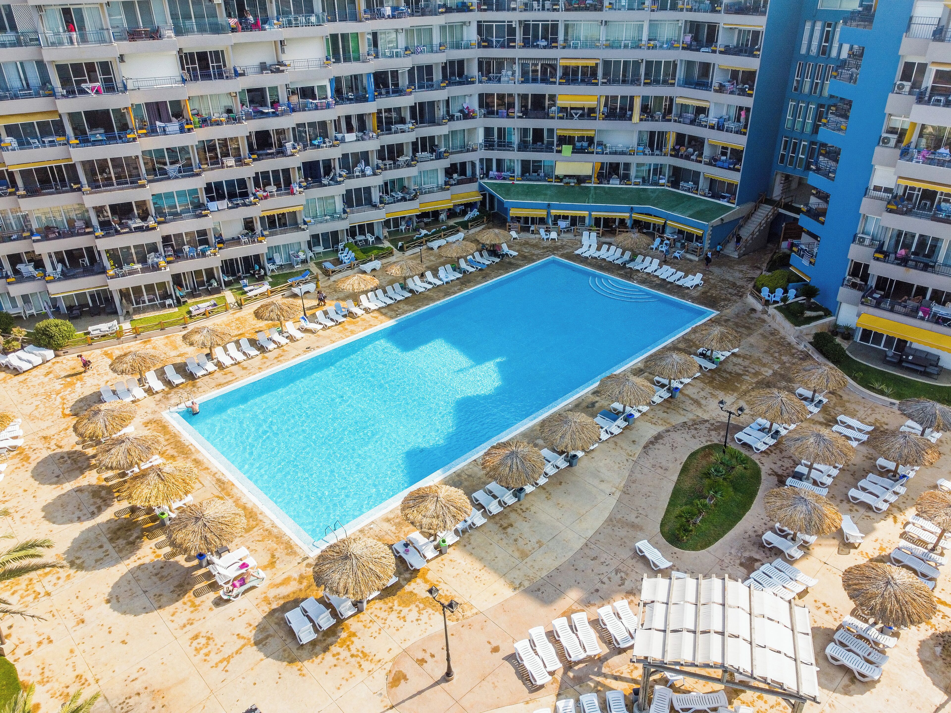 An aerial view of a hotel with multiple balconies, a central swimming pool, and sun loungers arranged around the pool.