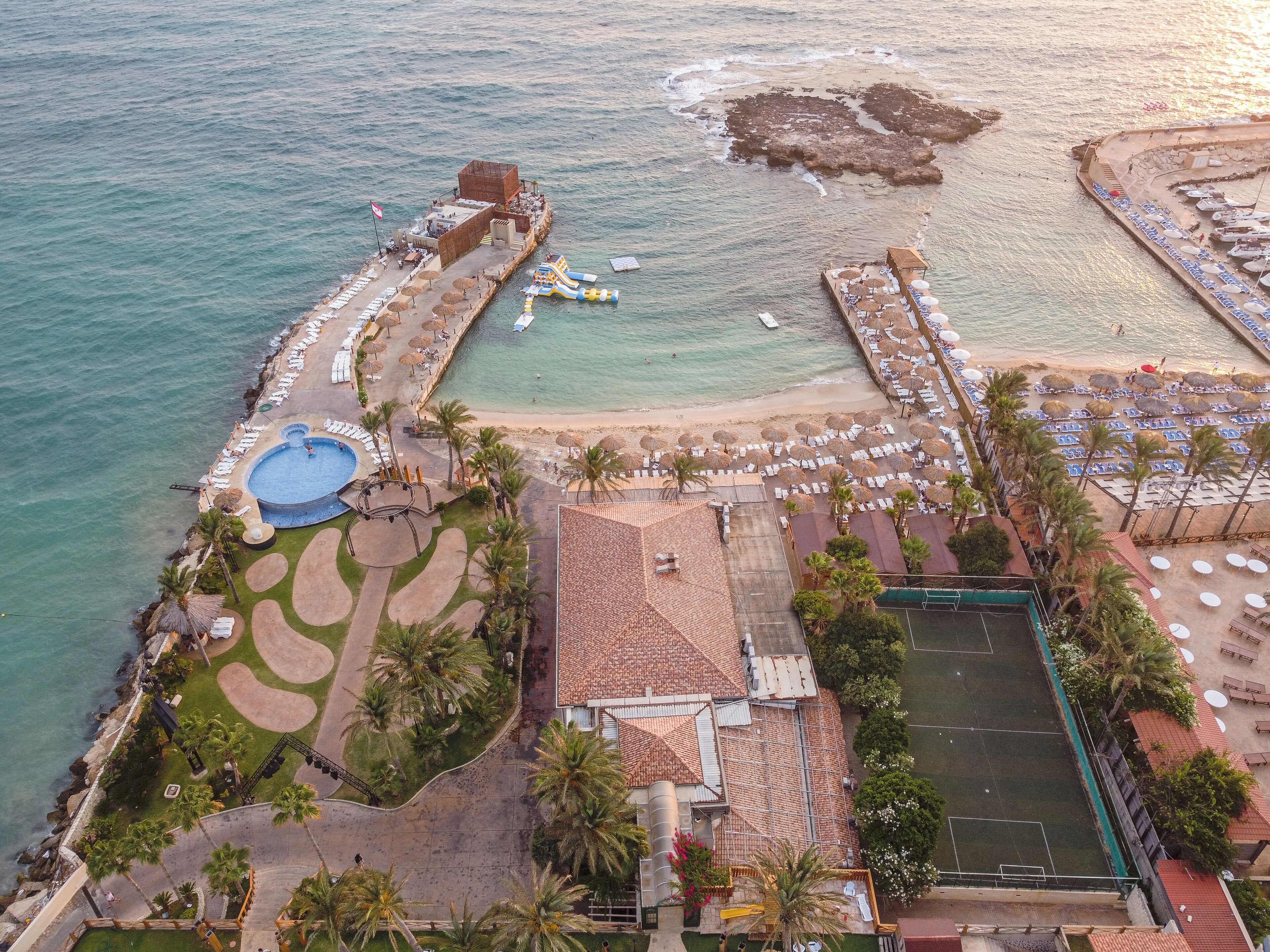 An aerial view of a coastal resort with a swimming pool, beach area with sun loungers, and a tennis court.