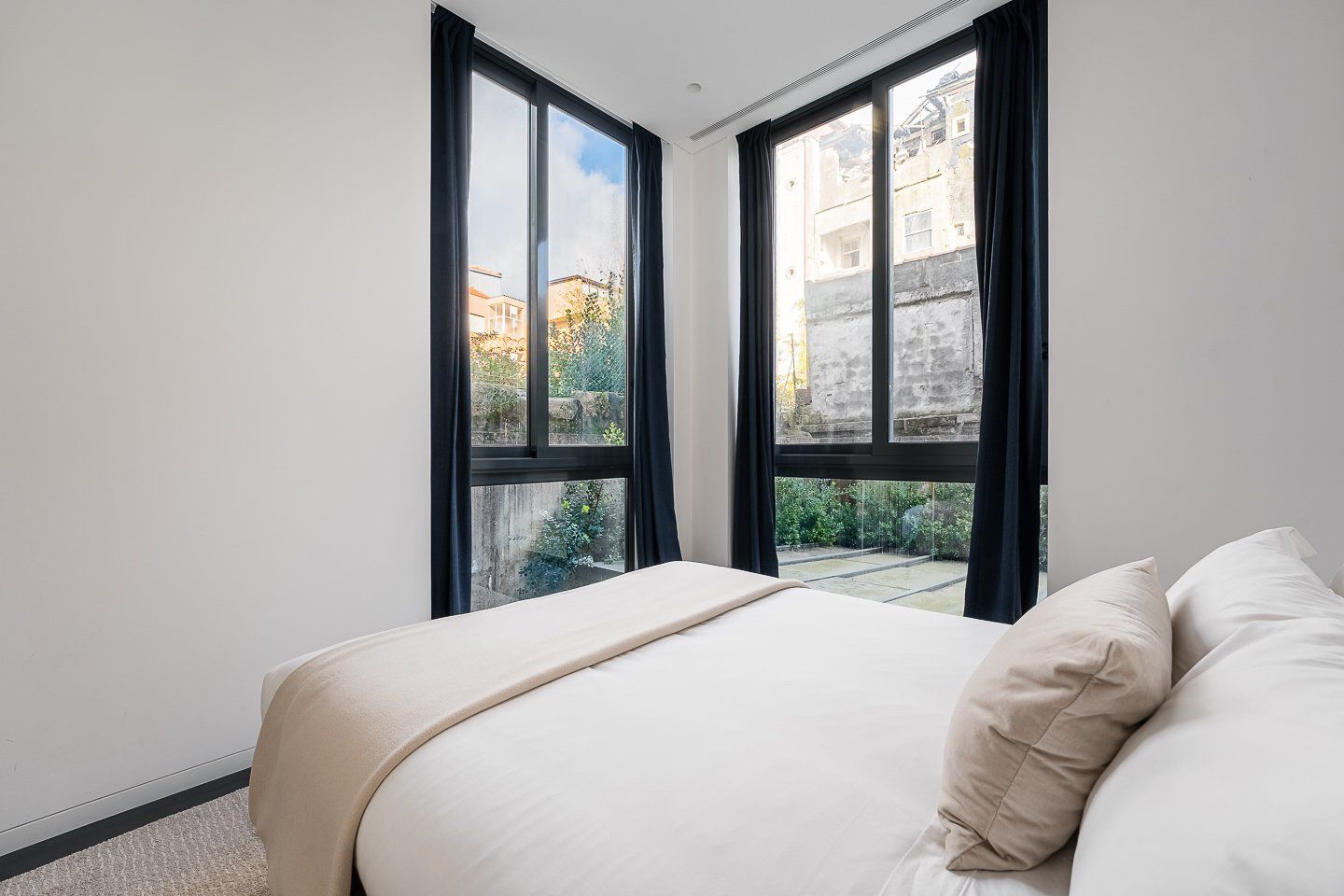 A bedroom with a bed, two windows, and a view of buildings and greenery outside.