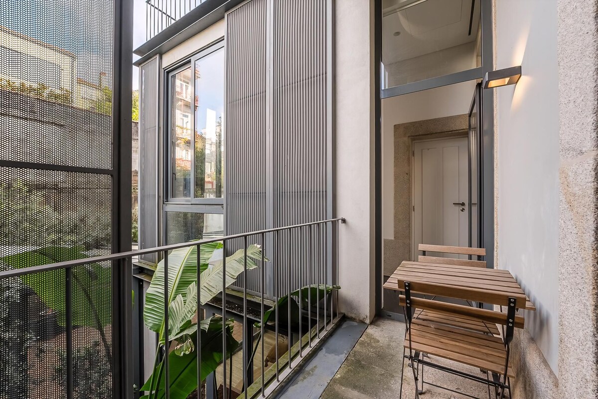 Een balkon met een metalen hekwerk, een houten tafel en stoelen, en uitzicht op gebouwen en groen.