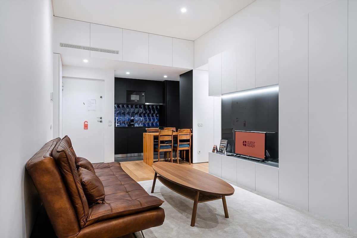 Una habitación de hotel moderna con sofá de cuero, una mesa de comedor de madera, zona de cocina con refrigerador y un ventanal grande.