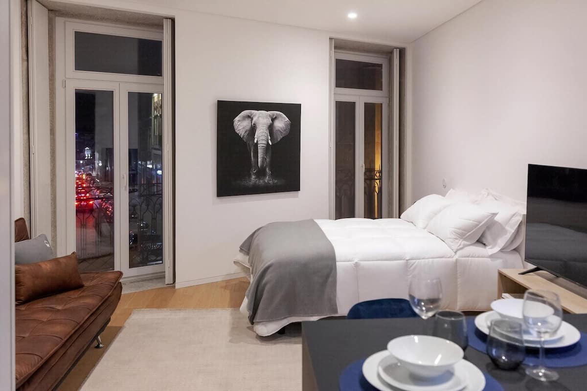 Habitación de hotel moderna con cama, una mesa de comedor con platos y vasos, un sofá, un cuadro de un elefante y vistas a la ciudad a través de amplias ventanas.