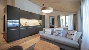Une cuisine moderne avec des meubles de couleur sombre et un îlot central, un canapé gris agrémenté de coussins à motifs et un plafond en bois.