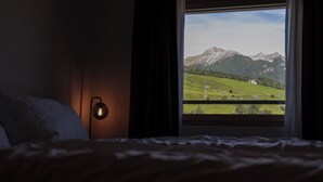 Une chambre à coucher avec un lit, une lampe de chevet et une fenêtre donnant sur une vallée verdoyante et des montagnes.