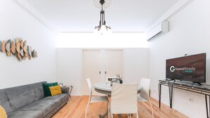 A modern living room with a grey sofa, a dining table with chairs, a chandelier, and a flat-screen TV. - Guestready - Gold Leaf (Porto)