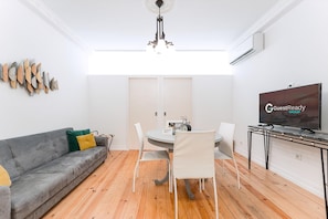 A modern living room with a grey sofa, a dining table with chairs, a chandelier, and a flat-screen TV. - Guestready - Gold Leaf (Porto)