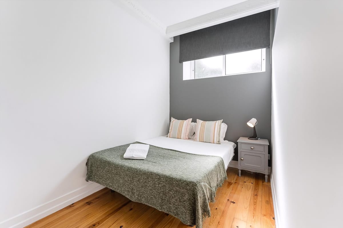 A bedroom with a bed, a nightstand, a window with a blind, and a wooden floor.