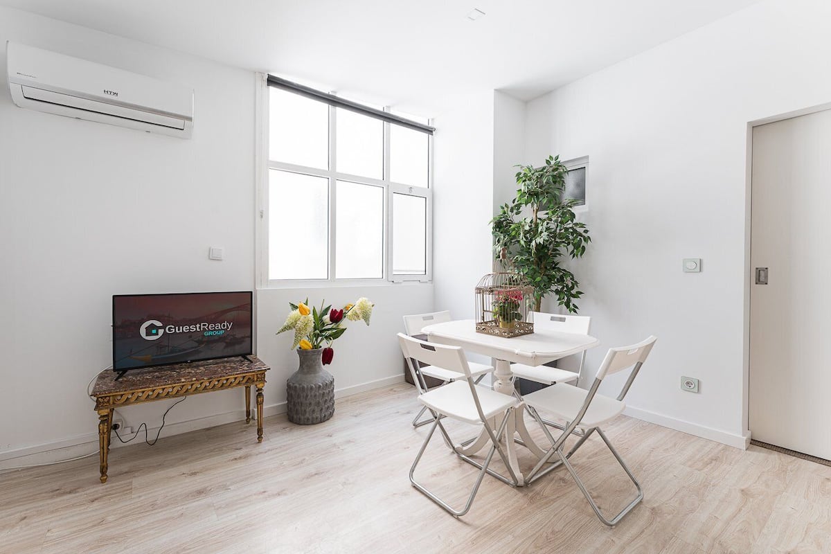 A modern living room with a flat-screen TV, a dining table with chairs, a potted plant, and a large window.