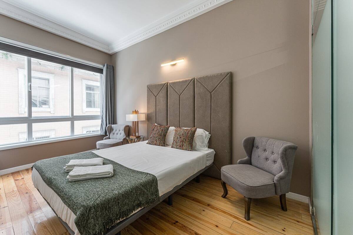 A hotel room with a large bed, a grey armchair, a bedside table with a lamp, and a view of buildings through the window.