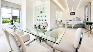 Un espace repas moderne avec une table en verre et des chaises blanches, un piano, et un coin cuisine doté d’un plan de travail et de meubles blancs.