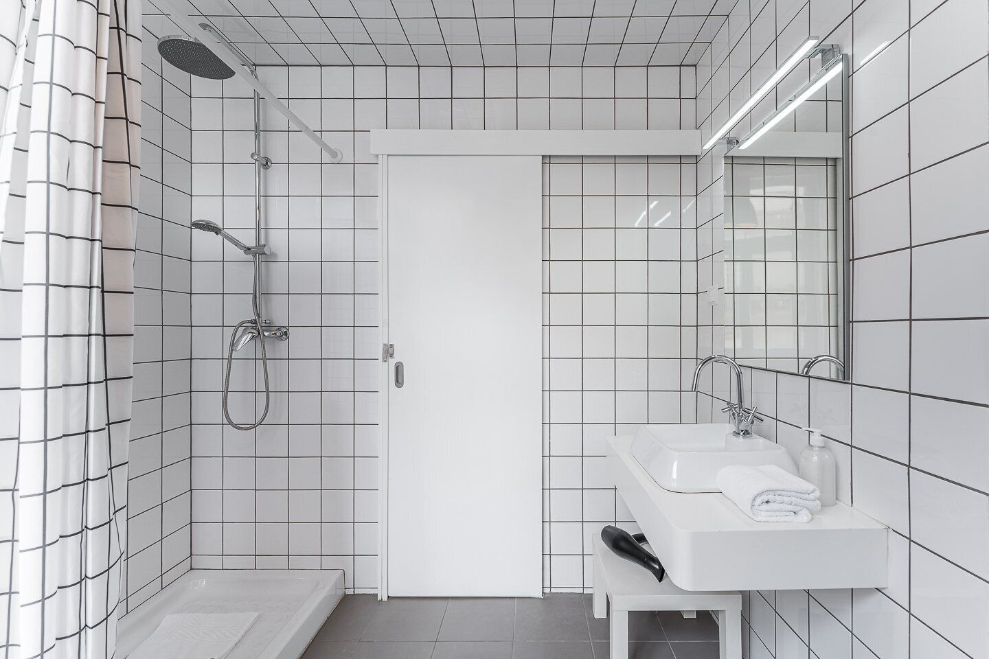 A modern bathroom with white tiled walls, a shower with a handheld showerhead, a sink with a mirror, and a towel on the sink.