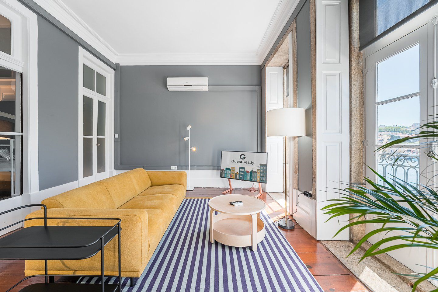 A modern living room with a mustard sofa, a small round table, a floor lamp, and a view of the city through large windows.