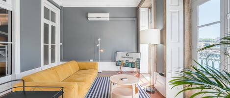 A modern living room with a mustard sofa, a small round table, a floor lamp, and a view of the city through large windows.