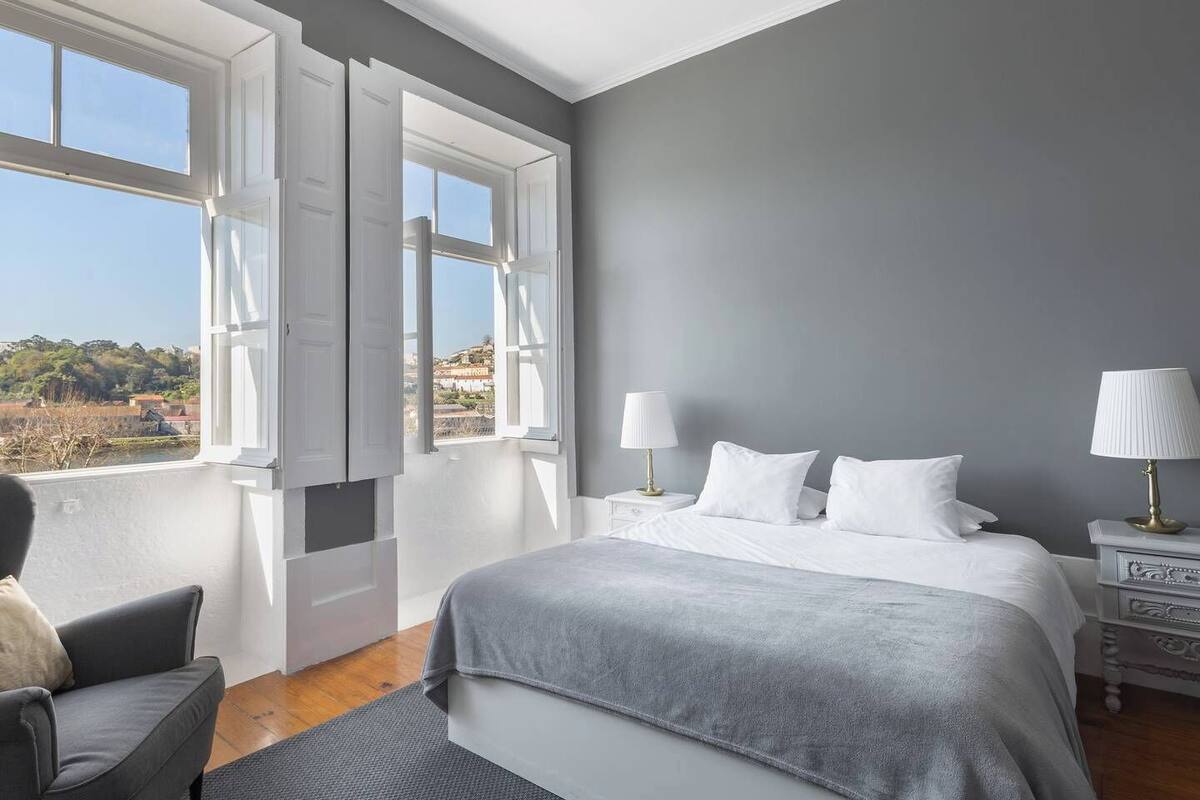 Une chambre dotée d’un grand lit, de deux tables de chevet avec des lampes, d’un fauteuil et offrant une vue sur l’extérieur grâce à des fenêtres ouvertes.