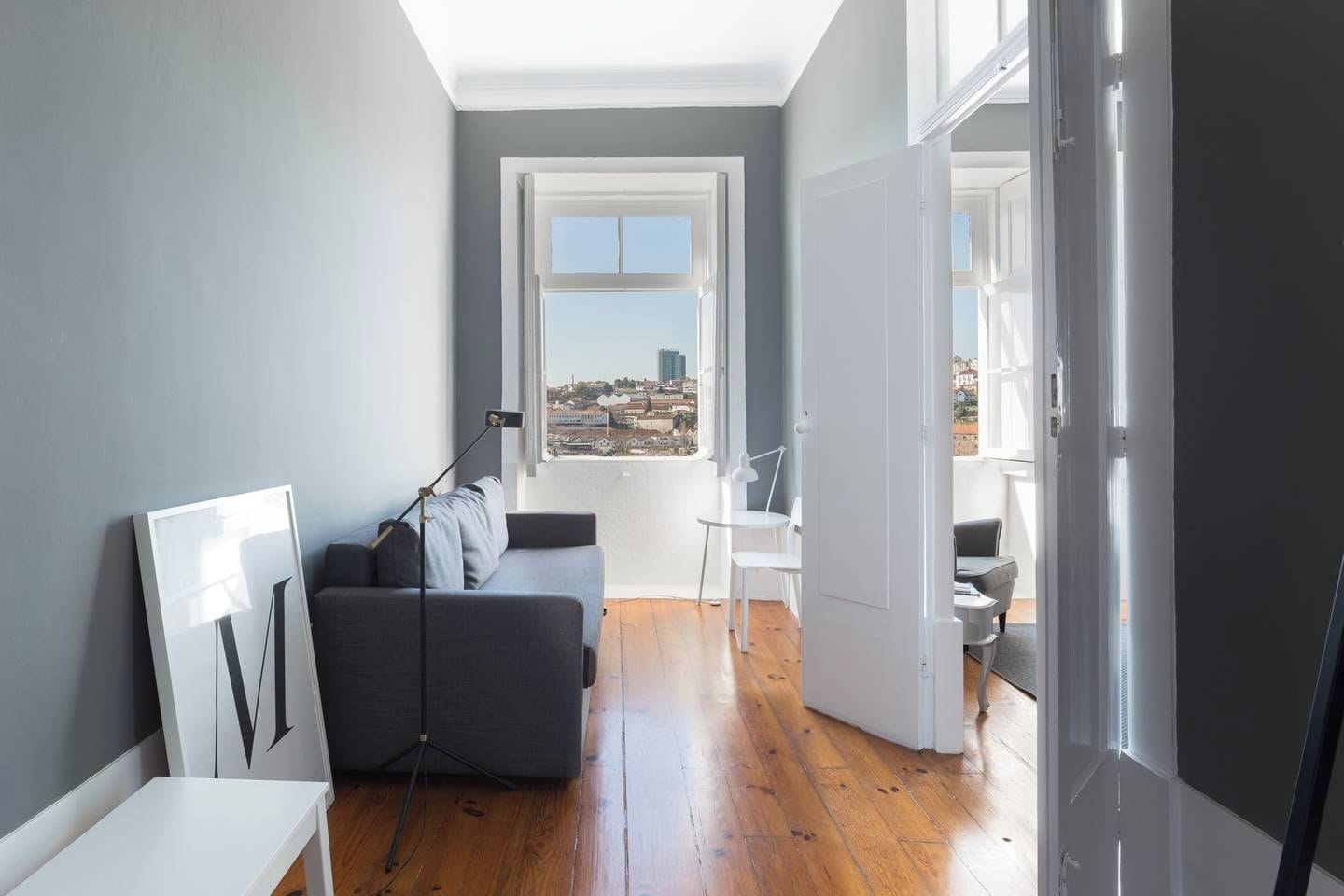 A modern living room with a grey sofa, a small white table, and a view of the city through a large window.