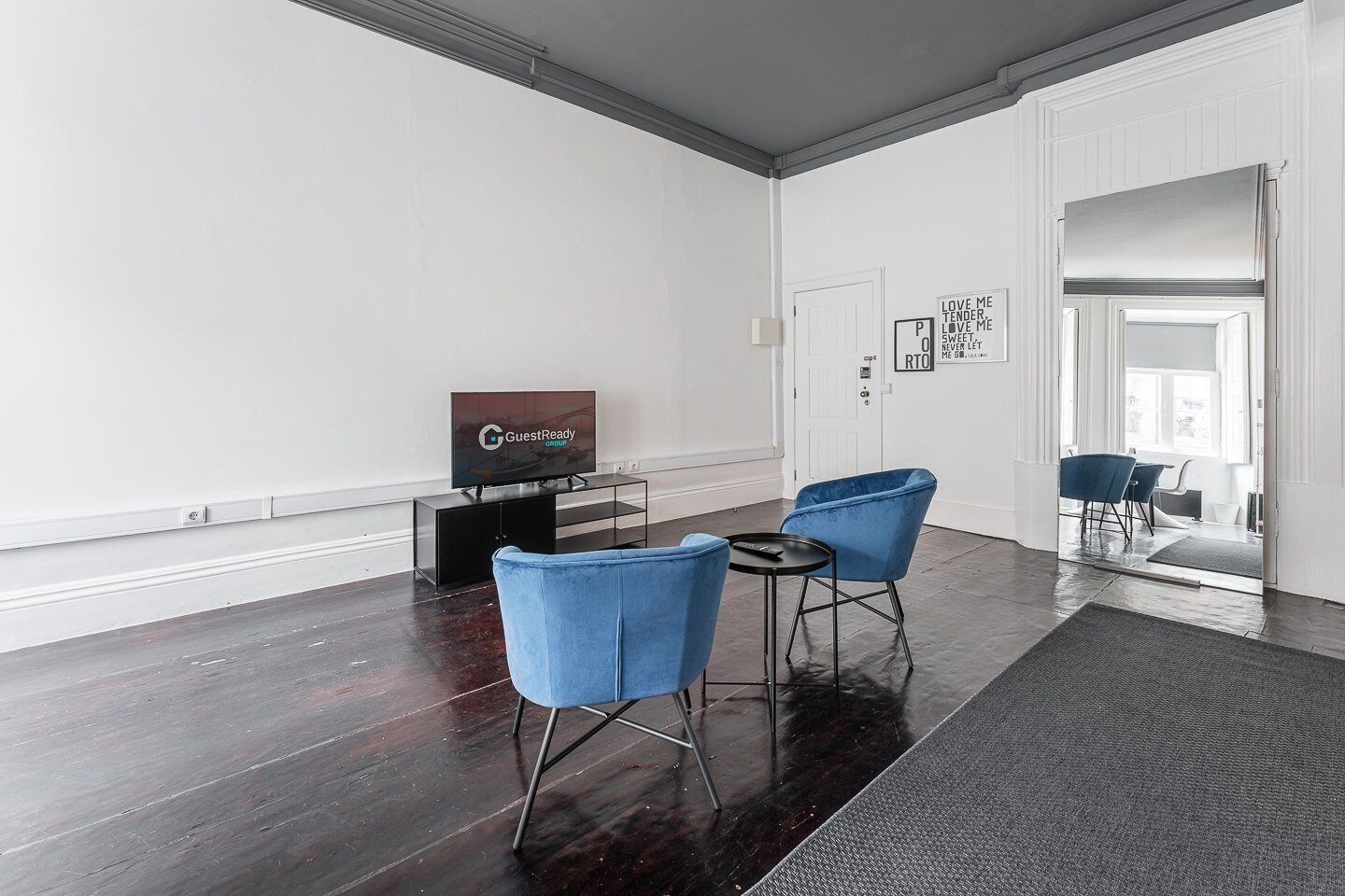 Ein modernes Wohnzimmer mit einem Flachbildfernseher, zwei blauen Sesseln, einem kleinen Tisch und einem dunklen Holzboden.