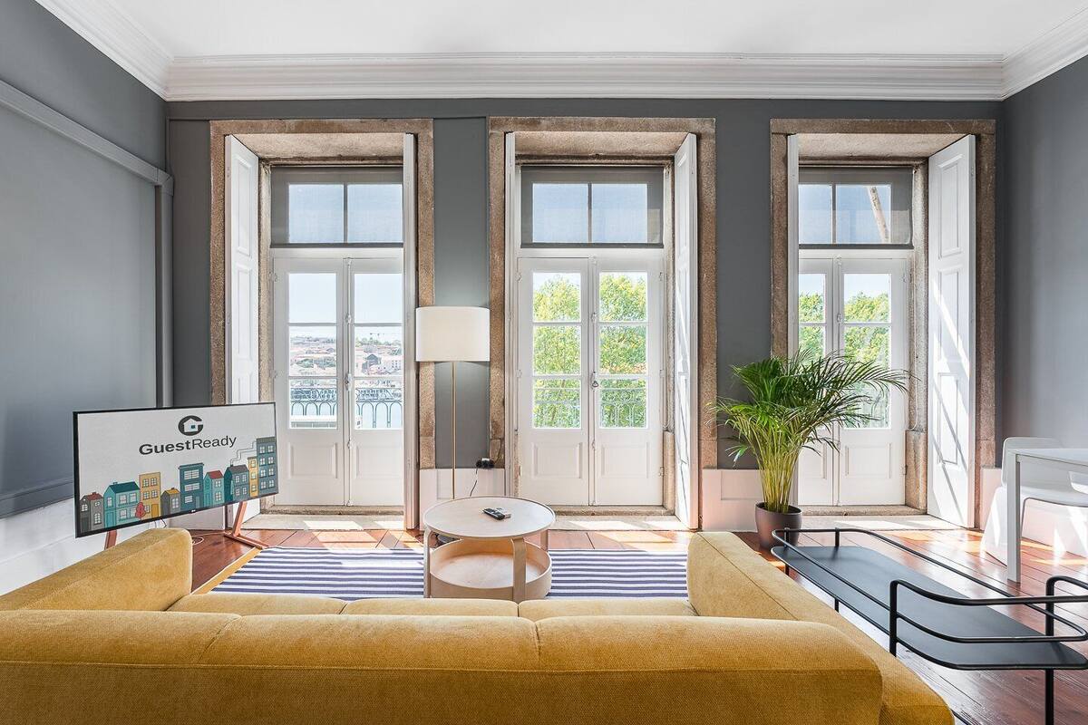 Un salon moderne avec un canapé, une petite table et une plante en pot. Il y a trois grandes fenêtres avec des rideaux, et un panneau accroché au mur.
