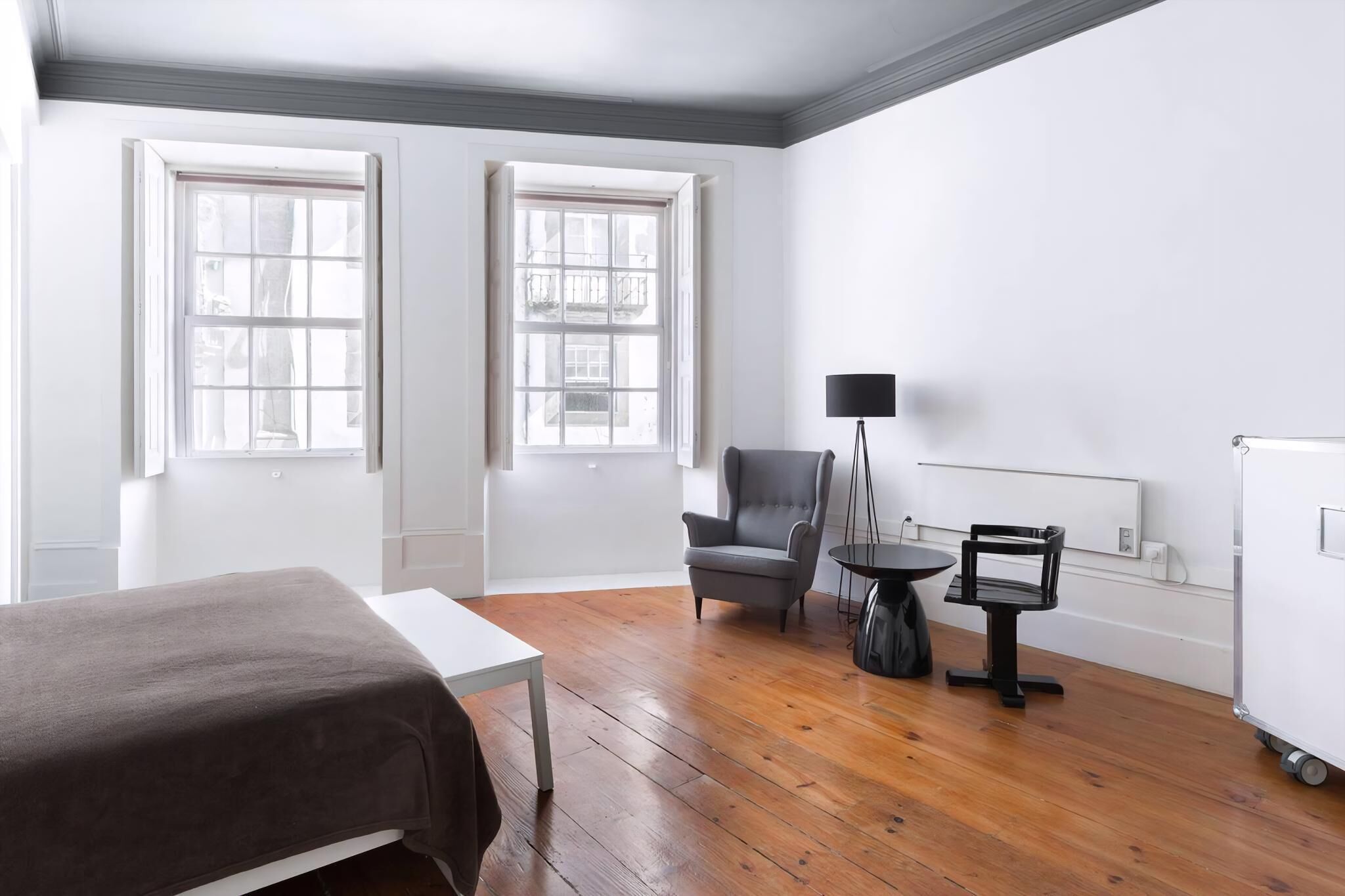 A hotel room with a bed, a chair, a small table, and a lamp. There are two windows and a radiator on the wall.