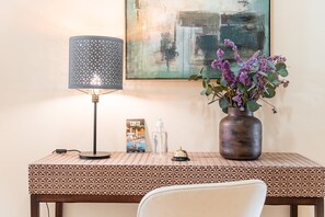A hotel desk with a patterned tablecloth, a lamp, a vase with flowers, a framed picture, a bottle, and a small bell. - Guestready - La Maison Aires Gouveia (Porto)